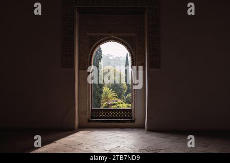 Lush trees and plants growing in garden seen through arched window in dark room of historic famous Alhambra fortress in Granada, Spain Foto Stock