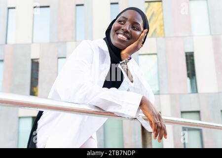 Dal basso, allegra donna d'affari afroamericana musulmana in hijab nero che tocca il mento e guarda lontano mentre si piega su una ringhiera in città Foto Stock