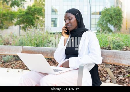 Una freelance musulmana concentrata in hijab nero che parla tramite smartphone e lavora a progetti in remoto su un computer portatile seduto sulla panchina Foto Stock