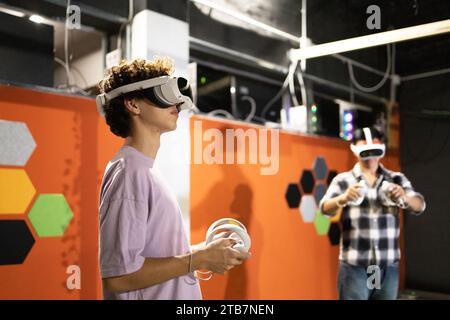 Due amici si trovano in un'area di gioco, completamente immersi in un mondo di realtà virtuale, indossando occhiali VR e tenendo in mano i controller di movimento. Foto Stock