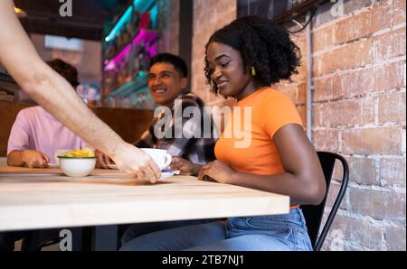 "Una giovane donna sorridente siede a un tavolo di legno in un bar murato di mattoni, condividendo un momento con gli amici." Foto Stock