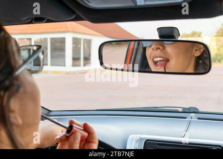 Affascinante giovane donna d'affari caucasica che guarda nello specchietto retrovisore e applica rossetto con spazzola mentre si siede in un'auto moderna Foto Stock