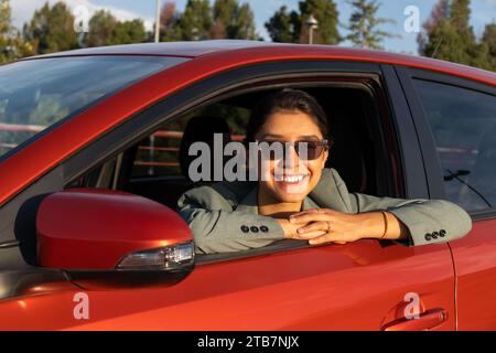 Ritratto di una giovane imprenditrice caucasica felice e attraente che indossa occhiali da sole e blazer seduto in auto arancione e guarda attraverso la finestra Foto Stock