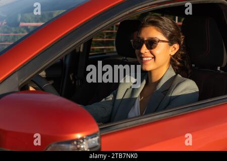 Allegra e attraente giovane imprenditrice caucasica che indossa occhiali da sole e blazer sorridente mentre guida una moderna automobile arancione in città Foto Stock