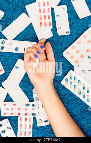 Dall'alto tagliata non riconoscibile mano di donna depressa che tiene varie pillole su uno sfondo blu sparse con bolle di farmaci vuote Foto Stock