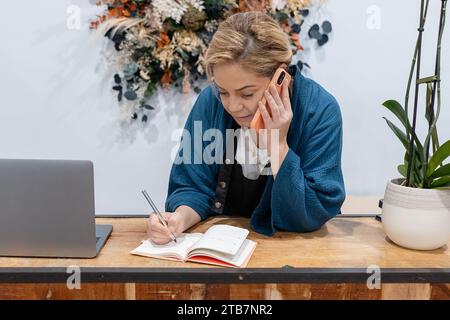 La donna si concentra sulla scrittura di appunti in un planner con un laptop su una scrivania di legno mentre parla al telefono cellulare Foto Stock