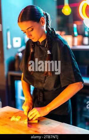 Giovane cuoca con trecce in uniforme nera con un ingrediente da taglio focalizzato con coltello sul tagliere mentre prepari il pasto a Illuminated Foto Stock