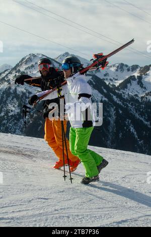 Un paio di sciatori che trasportano le loro attrezzature attraversano una pista innevata con le maestose Alpi svizzere sullo sfondo Foto Stock