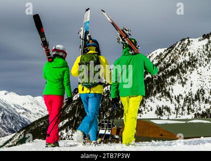 Tre sciatori in una vivace attrezzatura che trasportano sci su uno sfondo innevato. Foto Stock