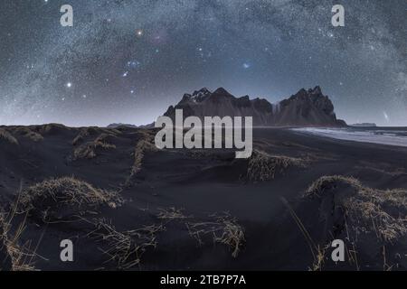 Un'ampia via Lattea brilla su un aspro paesaggio costiero con ciuffi di erba secca che adornano le dune di sabbia nera Foto Stock