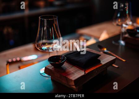 Un ambiente intimo di un ristorante di sushi con un tavolo in legno, eleganti bicchieri e un piatto di pietra in attesa di una cucina deliziosa. Foto Stock