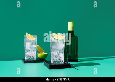 Composizione artistica composta da due bicchieri con spicchi di gin tonic e lime, insieme a una bottiglia di whisky scura, su uno sfondo verde vibrante Foto Stock