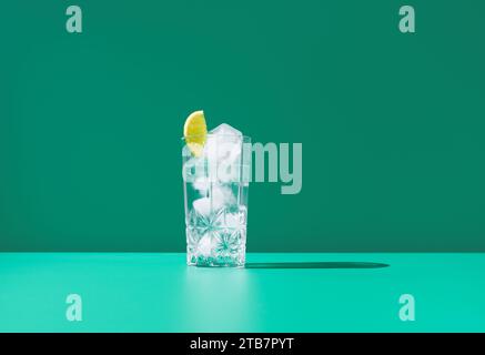 Un vetro trasparente pieno di gin tonic e ricoperto da una fetta di lime si trova su uno sfondo verde vibrante, creando un aspetto fresco e fresco. Foto Stock