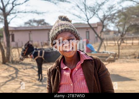 felice donna africana del villaggio, che ride per strada, strada sterrata con un carretto d'asino sullo sfondo Foto Stock