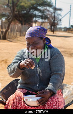 villager, una vecchia donna africana in una carriola che mangia del cibo tradizionale e ride di una buona battuta Foto Stock