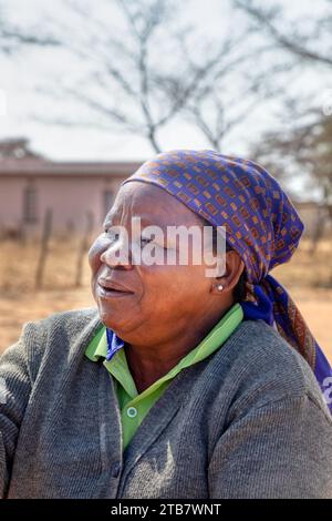 villager , colpo di testa di vecchia donna africana con sciarpa blu, casa e strada sterrata sullo sfondo Foto Stock