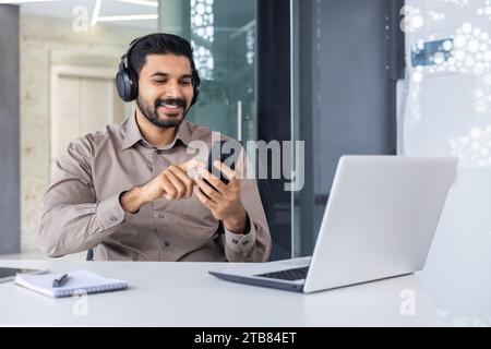 Uomo indù soddisfatto di successo sul posto di lavoro all'interno dell'ufficio, uomo con telefono in mano e cuffie che guarda video online e ascolta musica audio p Foto Stock
