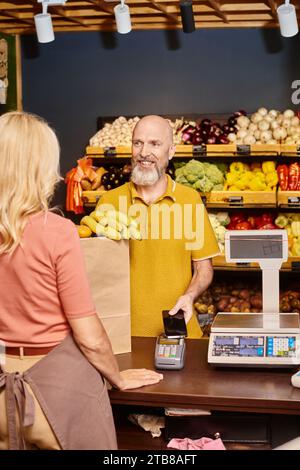 concentrati su un uomo dalla barba allegra che paga con il telefono per comprare generi alimentari freschi e sorridere da una venditrice Foto Stock