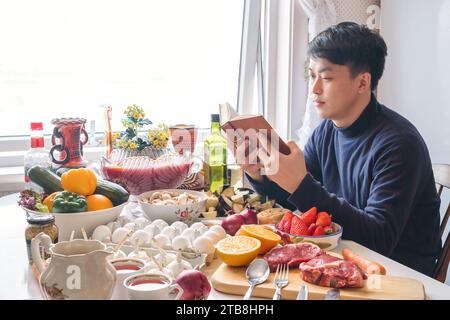 Giovane asiatico che legge un libro e cena da solo al tavolo da pranzo, festeggia cibo e bevande nel giorno del Ringraziamento in inverno Foto Stock