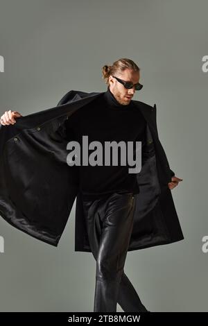 ritratto di un bell'uomo barbuto con occhiali da sole alla moda e abbigliamento nero che cammina su sfondo grigio Foto Stock