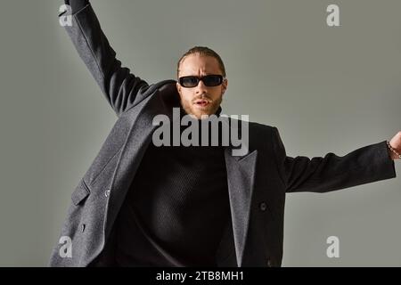 ritratto di un uomo barbuto con occhiali da sole alla moda e abbigliamento nero in posa con le mani alzate Foto Stock