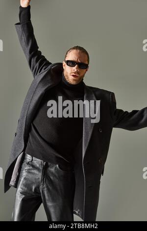 ritratto di un uomo barbuto con occhiali da sole alla moda e abbigliamento nero in posa con le mani alzate Foto Stock