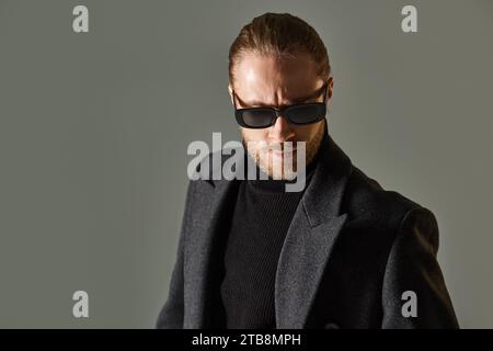ritratto di un uomo barbuto con occhiali da sole alla moda e abbigliamento nero in posa su sfondo grigio Foto Stock