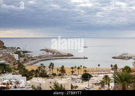 Porto Rico, Spagna - 26 novembre 2023: Spiaggia turistica con ombrelloni e porto con barche a Puerto Rico de Gran Canaria, Las Palmas, Spanje Foto Stock