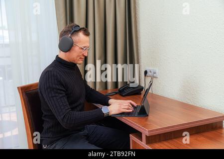 Uomo asiatico freelance che indossa occhiali e cuffie che lavora a casa e utilizza un tablet e si siede accanto al tavolo di legno Foto Stock