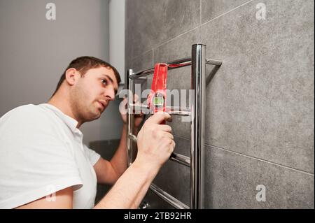 Idraulico maschio che controlla portasciugamani riscaldato elettrico con righello di livella a bolla d'aria in bagno. Uomo che installa un termoasciugamani in acciaio inox nell'appartamento in fase di ristrutturazione. Foto Stock