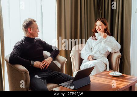 Primo piano di una coppia asiatica seduto su una sedia durante la luna di miele nella camera d'albergo e parlando. Uomini che guardano moglie e donna che bevono tè dalla tazza Foto Stock