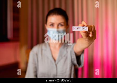 Primo piano di una donna caucasica in maschera protettiva che tiene in mano un risultato positivo del dispositivo di prova. Giovane donna che mostra il suo test rapido positivo al Covid-19 Foto Stock