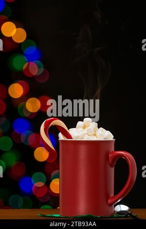 Una grande tazza rossa di cioccolata calda fumante condita con mini marshmallow e una canna da zucchero contro il bokeh di un albero di Natale Foto Stock