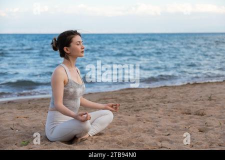 Giovane bella donna che pratica yoga all'aperto seduto sulla riva di un lago in posa di loto, rilassata, respirata profondamente e meditata. Seduto sulla sabbia vicino W. Foto Stock