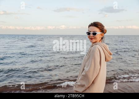 Giovane donna sottile con abiti in avorio e occhiali da sole bianchi in piedi sulla spiaggia incrociate le mani vicino al lago guardando la fotocamera godendo di una splendida vista. Esterno Foto Stock