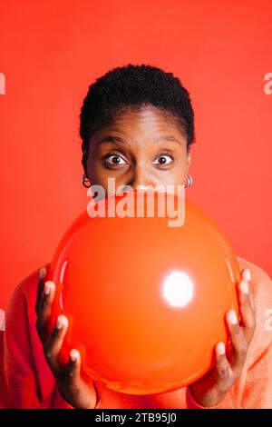 Ritratto di una giovane donna felice che soffia un palloncino rosso davanti a uno sfondo rosso in uno studio. Donna con capelli corti che gonfia un palloncino rosso. Foto Stock