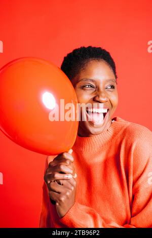 Ritratto di una giovane donna felice che soffia un palloncino rosso davanti a uno sfondo rosso in uno studio. Donna con capelli corti che gonfia un palloncino rosso. Foto Stock