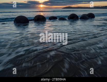 Marea in arrivo all'alba con i massi di Moeraki nel surf lungo un tratto di spiaggia di Koekohe sull'Isola Sud della nuova Zelanda Foto Stock