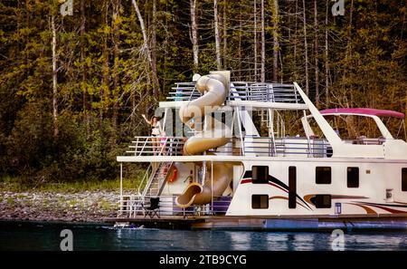 Una famiglia che si gode una vacanza in casa galleggiante mentre parcheggia sulla riva del lago Shuswap con mamma e figlia che saltano dal ponte a poppa del boa... Foto Stock