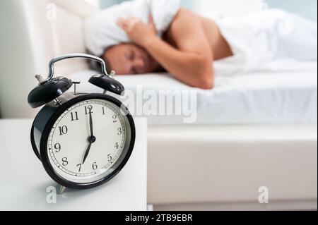 Sveglia nera sul comodino vicino a un uomo sfocato che dorme sul letto con un cuscino sulla testa. Alzarsi presto per andare al lavoro Foto Stock