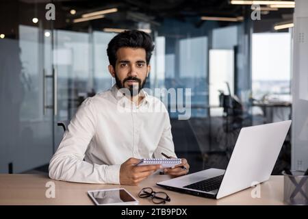 Ritratto di un giovane uomo d'affari indiano che lavora in ufficio a un tavolo con un laptop, che tiene un notebook, guarda seriamente la fotocamera. Foto Stock