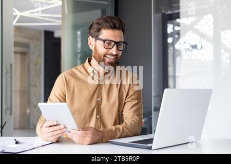 Ritratto di un giovane architetto, designer che lavora in ufficio con un laptop, parla durante una videochiamata, tiene un tablet tra le mani. Foto Stock