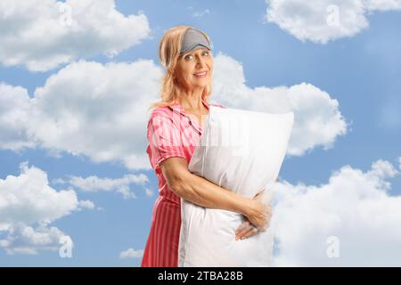 Donna in pigiama e una maschera addormentata che abbraccia un cuscino, nuvole e cielo blu come sfondo Foto Stock