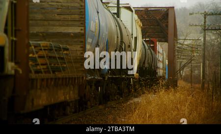 Un treno merci in movimento lento si avvicina e attraversa un ponte arrugginito in un paesaggio rustico e desolato del midwest Foto Stock