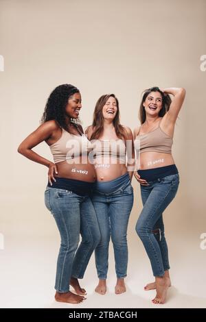 Gruppo multiculturale di donne incinte nel terzo trimestre in piedi insieme, sorridendo e mostrando i loro dossi. Amici in jeans e reggiseni Foto Stock