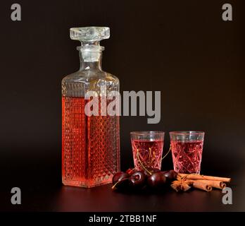 Liquore alla ciliegia con spezie, forte alcol fatto in casa in un decanter di cristallo e due bicchieri su sfondo nero, accanto a bastoncini di anice e cannella. CL Foto Stock