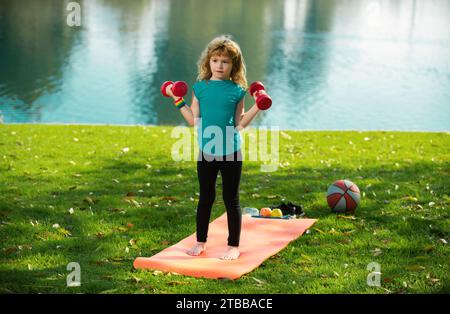 Simpatico bambino che tira i muscoli delle braccia con il manubrio all'aperto nel parco estivo. Fitness per bambini con manubri Foto Stock
