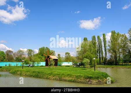 Piccolo lago con un mulino a legna e un'isola dal Parco Chindiei (Parcul Chindiei) a Targoviste, Romania, in una soleggiata giornata primaverile con nuvole bianche e blu Foto Stock