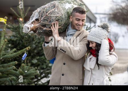 Un giovane che porta un albero di Natale avvolto, comprato al mercato insieme a sua moglie. La coppia felice ha comprato un albero di Natale. Foto Stock