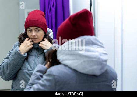 Una donna di mezza età sta provando una giacca e un cappello eleganti in un camerino presso un punto di prelievo. Sta esaminando il vestito e guardando allo specchio Foto Stock